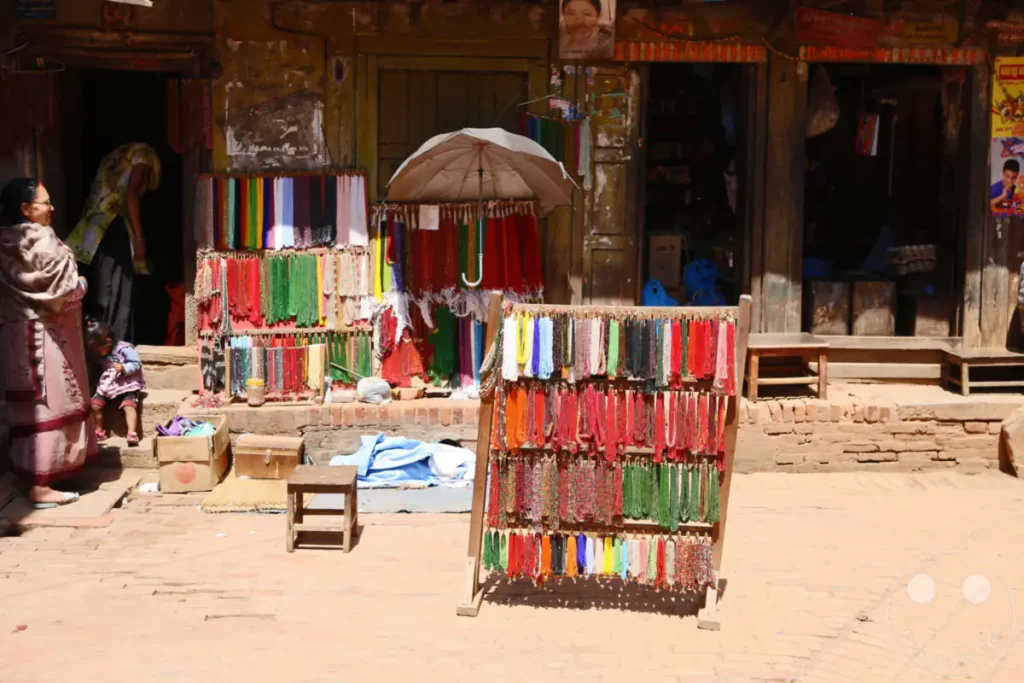 Ein farbenfroher Marktstand in Bhaktapur, Nepal, präsentiert eine Vielfalt an Perlenketten in zahlreichen Farben und Designs. Die Szene fängt die lebhafte Atmosphäre eines lokalen Marktes ein, mit traditioneller Kleidung und einem Einblick in die Geschäftigkeit.