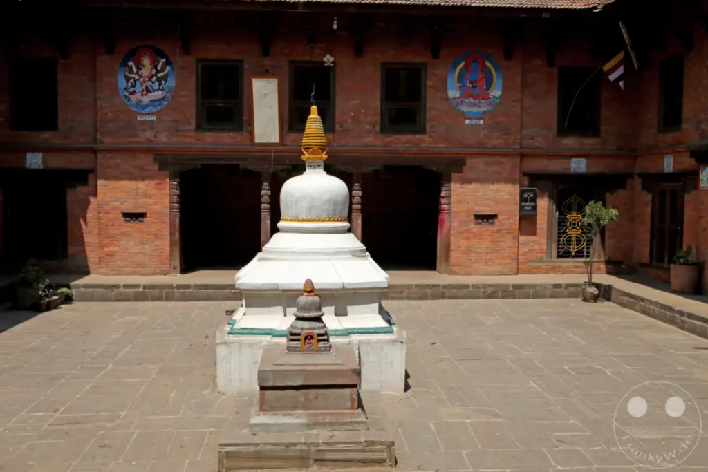 Ein Innenhof in Bhaktapur, Nepal, zeigt eine weiße Stupa im Vordergrund. Die umliegenden Backsteinmauern sind mit religiösen Wandmalereien und Fenstern verziert. Der Boden ist mit rechteckigen Steinplatten ausgelegt, die eine ruhige Atmosphäre schaffen.