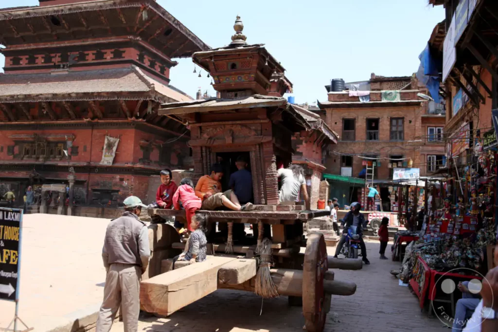 Ein großer hölzerner Tempelwagen steht auf einem Platz in Bhaktapur, Nepal. Kinder sitzen darauf, während im Hintergrund Geschäfte und Gebäude aus Ziegeln zu sehen sind. Die Szene zeigt das alltägliche Leben in der historischen Stadt.