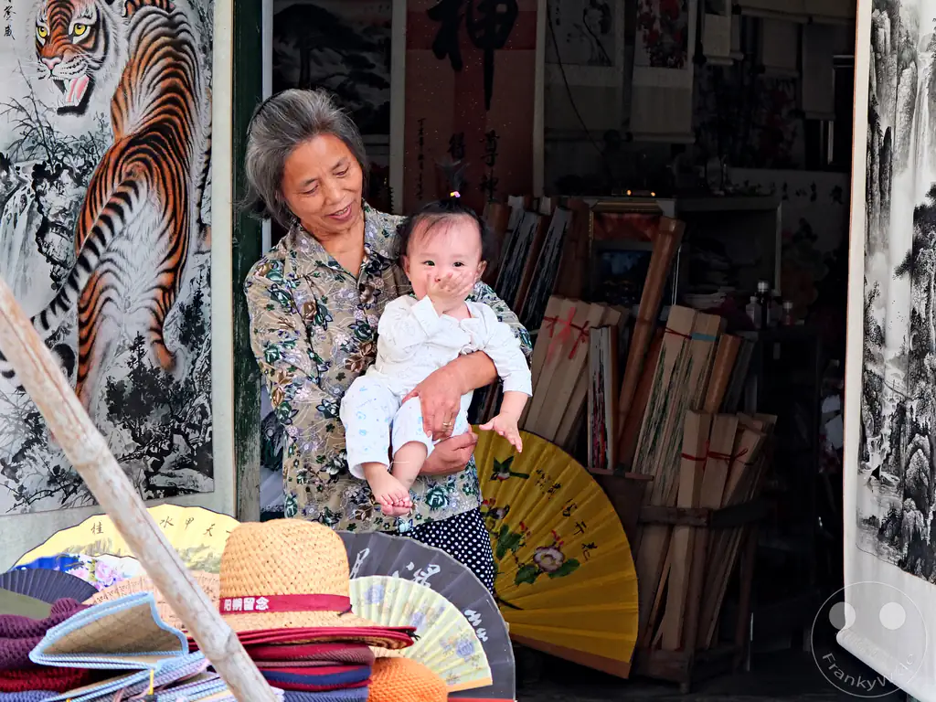 China - Yangshou - older woman smiling warmly while holding a young baby in her arms in front of an art or souvenir shop