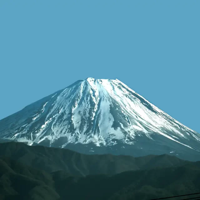 Der majestätische Berg Fuji mit seiner charakteristischen, tief verschneiten Vulkankegel-Spitze erhebt sich hinter einer dunklen Hügelkette vor einem klaren, hellblauen Himmel in Japan.