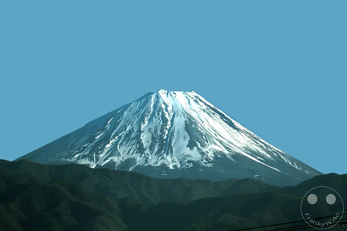 Der majestätische Berg Fuji mit seiner charakteristischen, tief verschneiten Vulkankegel-Spitze erhebt sich hinter einer dunklen Hügelkette vor einem klaren, hellblauen Himmel in Japan.