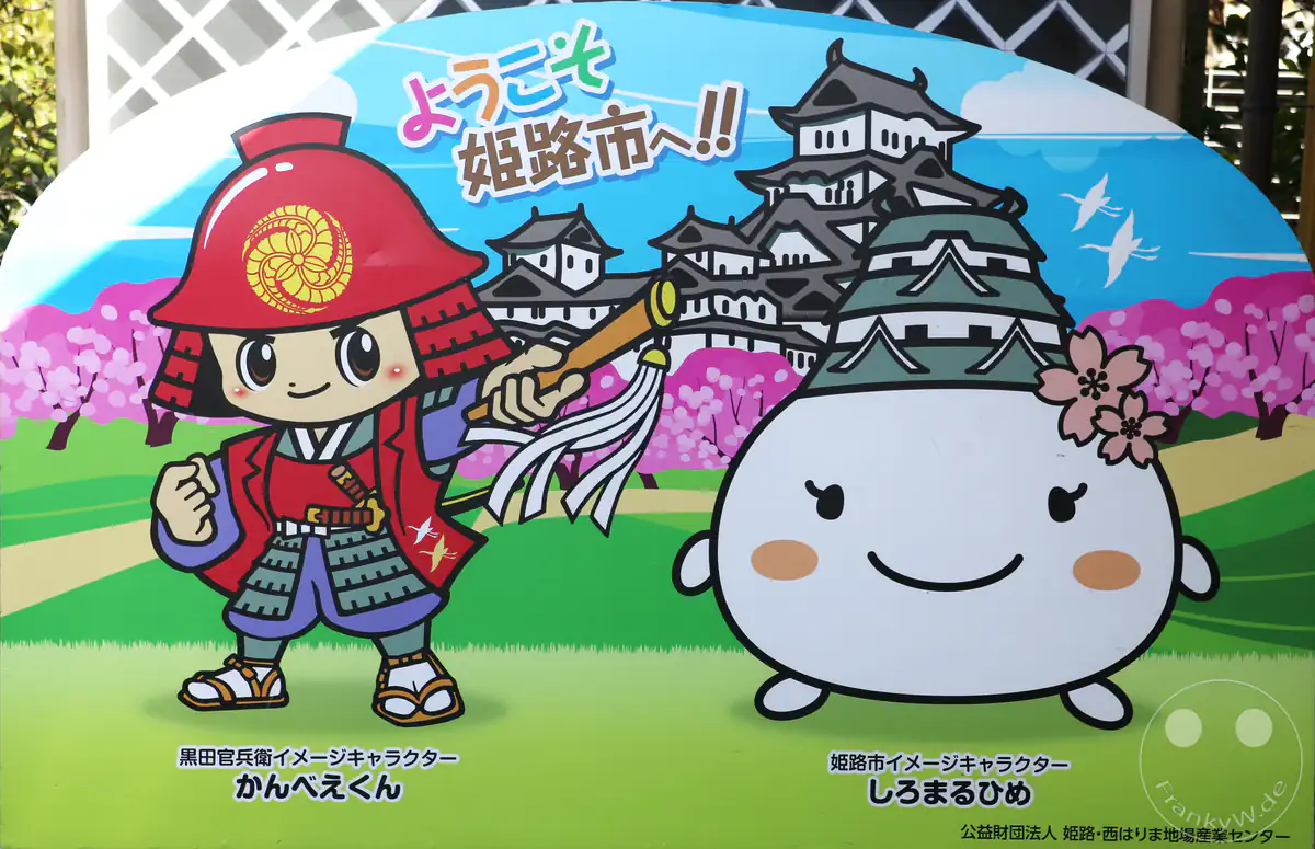 Eine bunte Willkommenstafel in Himeji zeigt die Maskottchen Shiromaruhime und Kanbee-kun vor der Kulisse der berühmten weißen Burg inmitten von blühenden Kirschbäumen.