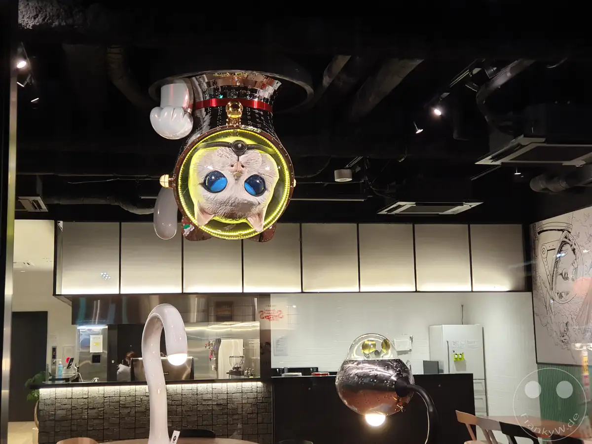 Modernes Café und Co-Working-Space mit großem Holztisch, stylischen Design-Möbeln und einer futuristischen Astronauten-Katzen-Skulptur als kreative Deckenbeleuchtung im japanischen Stil.