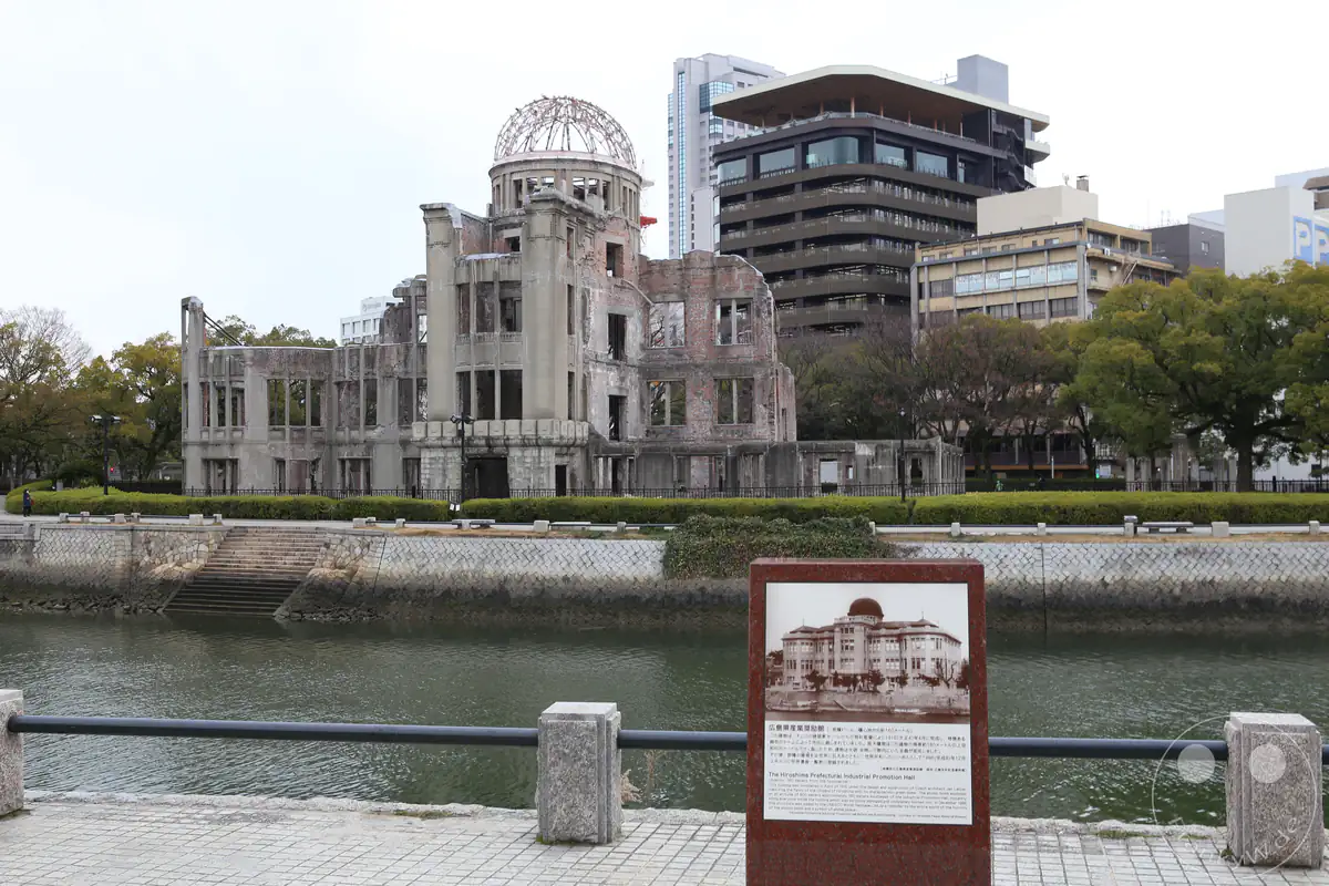 Der Atombombendom in Hiroshima steht als mahnendes Friedensdenkmal am Flussufer, ergänzt durch eine historische Informationstafel mit einer Fotografie des Gebäudes vor der Zerstörung.