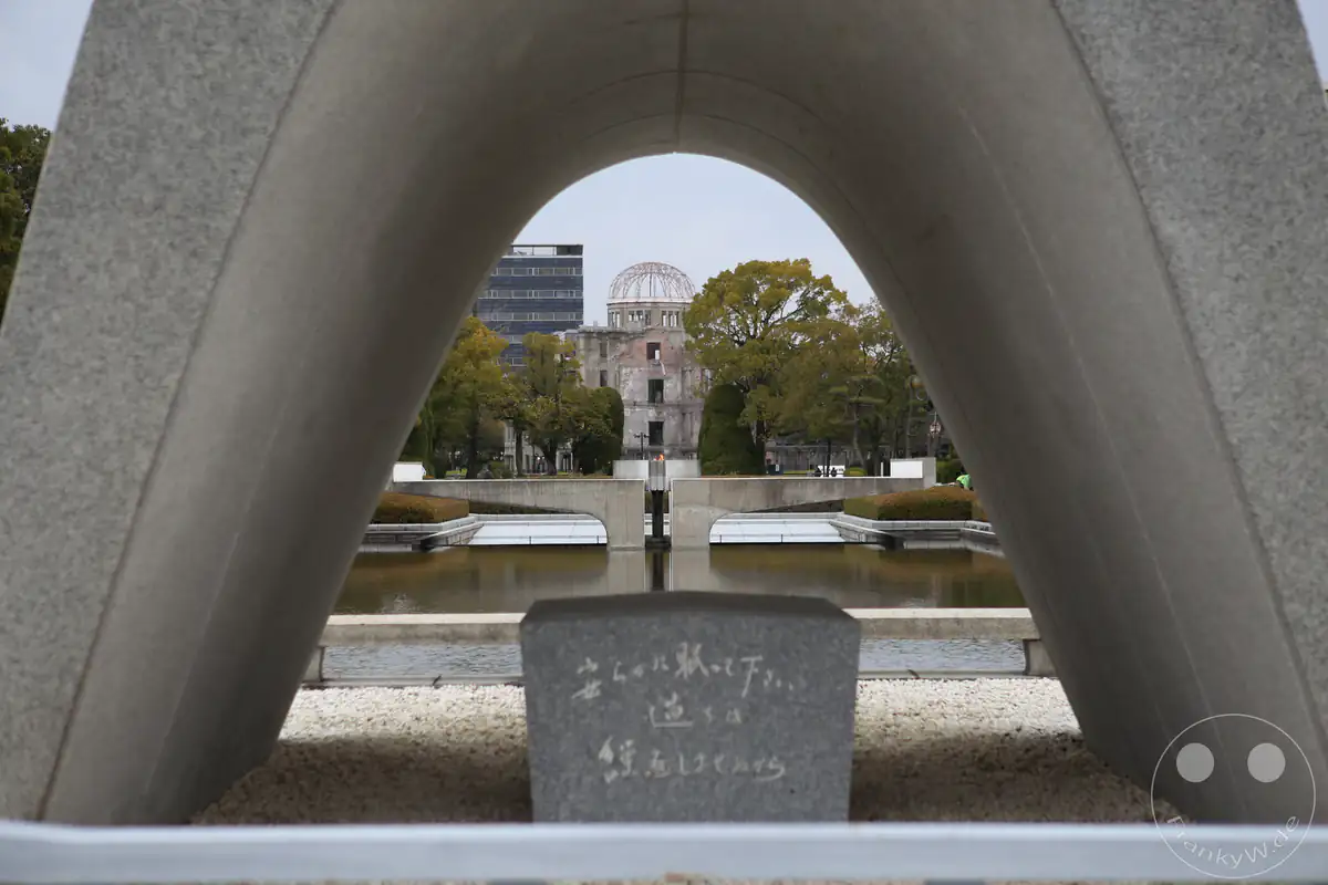 Blick durch das bogenförmige Beton-Kenotaph auf die Ewige Friedensflamme und den historischen Atombombendom im Peace Memorial Park in Hiroshima, Japan als symbolisches Mahnmal gegen Atomwaffen.