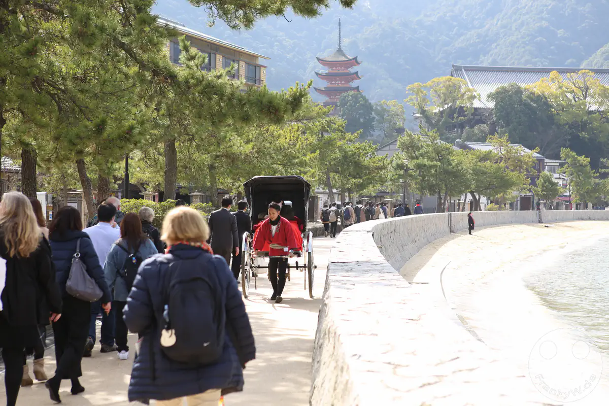 Touristen flanieren an der Uferpromenade auf der Insel Miyajima, während eine traditionelle Rikscha vorbeifährt und die fünfstöckige Gojunoto-Pagode im Hintergrund aufragt.
