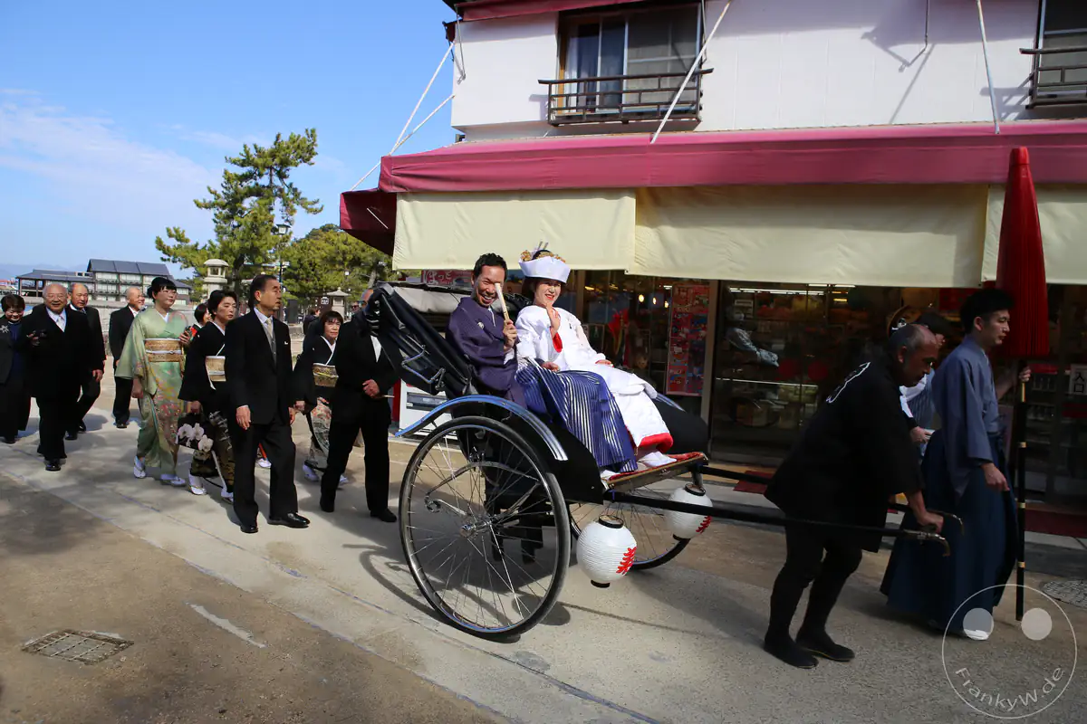 Ein japanisches Brautpaar in traditioneller Hochzeitskleidung fährt glücklich in einer Rikscha, gefolgt von einer Hochzeitsgesellschaft in feierlichen Kimonos und Anzügen auf Miyajima.