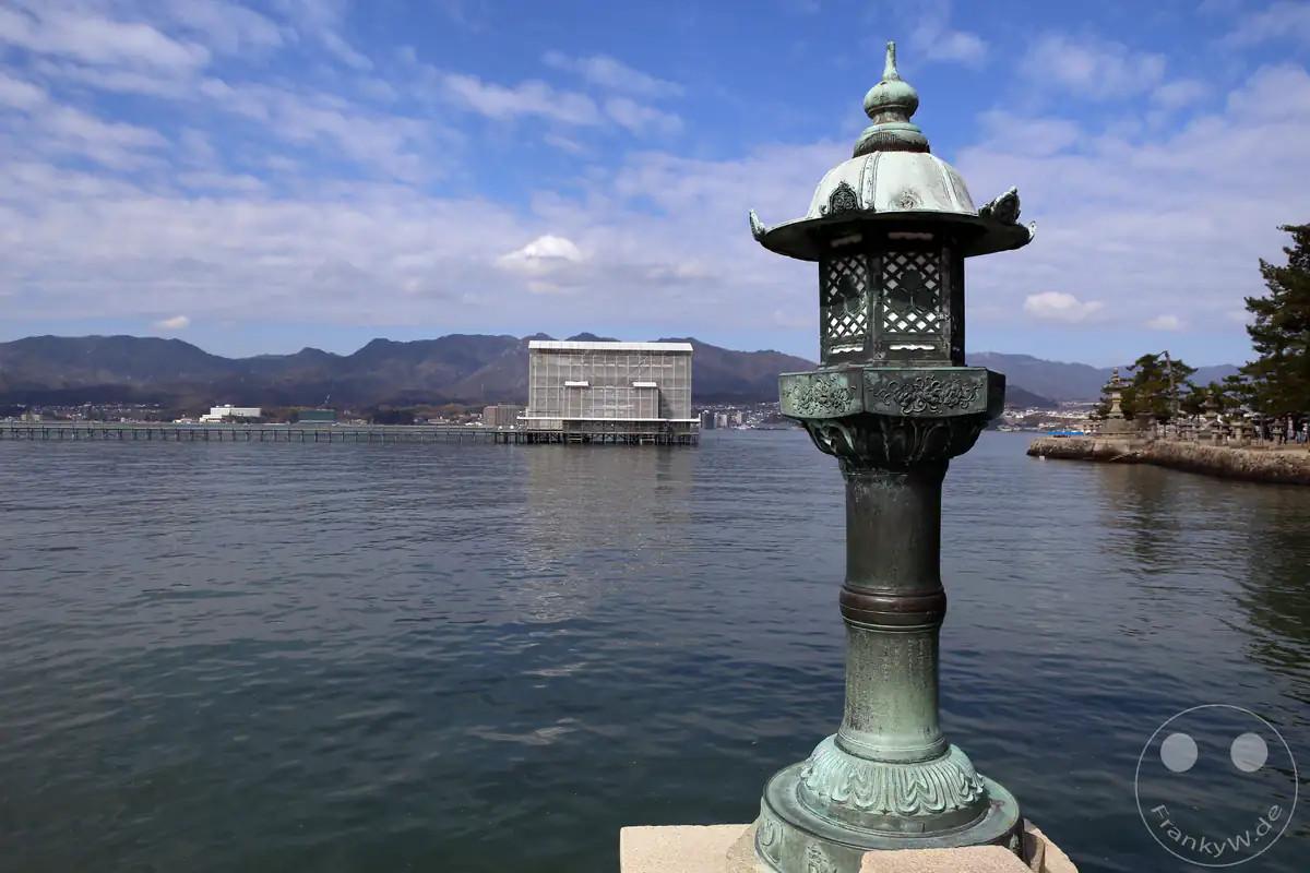 Eine traditionelle bronzene Steinlaterne steht im Vordergrund am Wasser, während im Hintergrund das verhüllte Torii-Tor des Itsukushima-Schreins auf Miyajima wegen Renovierungsarbeiten eingerüstet ist.