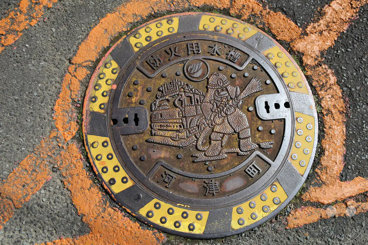 Ein reich verzierter, gusseiserner Hydrantendeckel in der Stadt Kawazu mit einem geprägten Motiv eines Feuerwehrmannes und eines Löschfahrzeugs, umgeben von einem gelb-schwarzen Warnring und einer leuchtend orangefarbenen Straßenmarkierung auf grauem Asphalt in der Präfektur Shizuoka, Japan.
