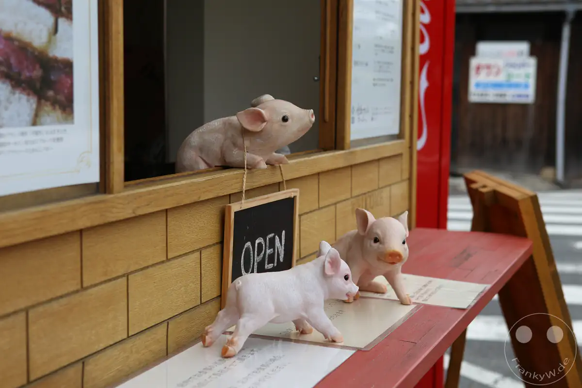 Drei kleine, lebensechte Schweinefiguren dekorieren die hölzerne Theke eines geöffneten Imbissstandes, ergänzt durch ein hängendes "Open"-Schild und ausliegende Speisekarten im Außenbereich.