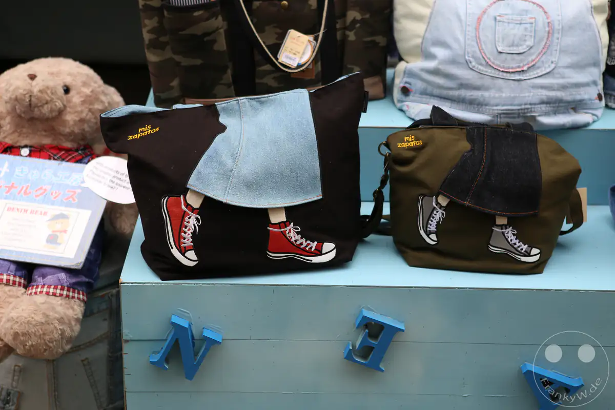 Zwei originelle Handtaschen der japanischen Marke mis zapatos, verziert mit Beinen in Jeansröcken und Sneakern, stehen dekorativ in einer Schaufensterauslage neben einem Plüschbären.