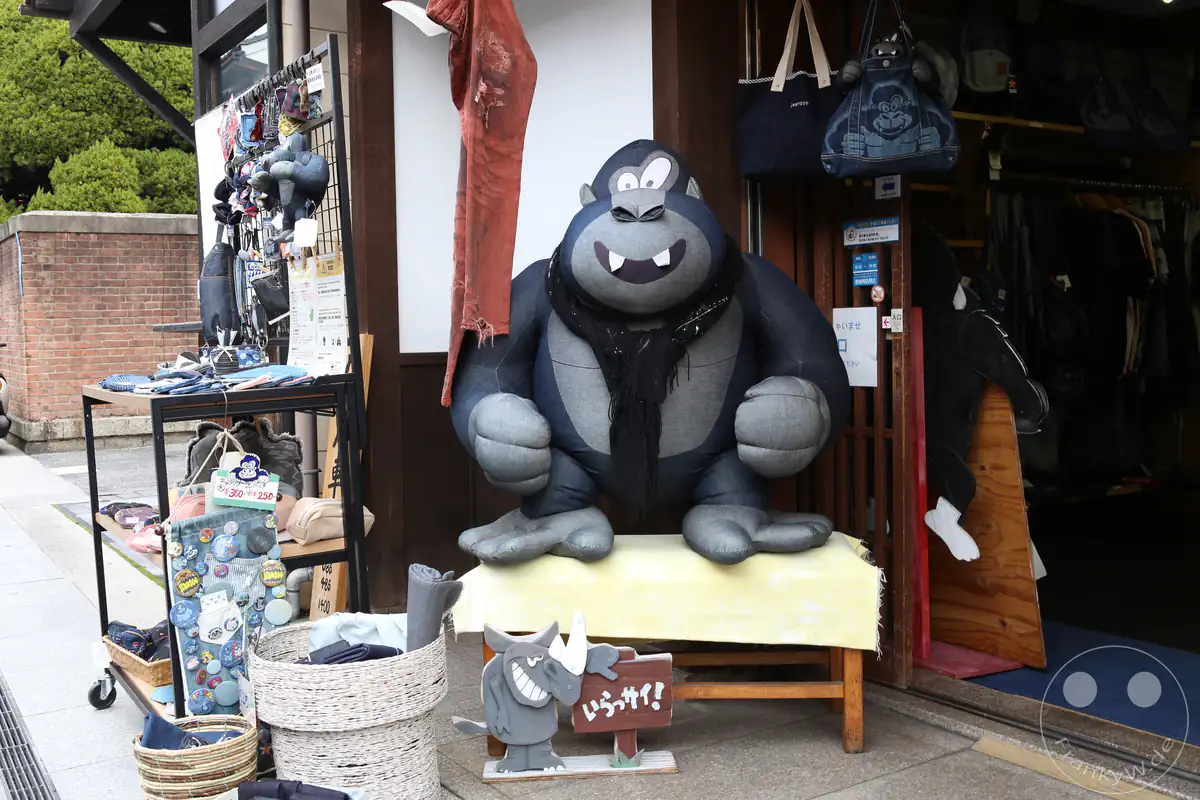 Eine große, aus Denim-Stoff gefertigte Gorilla-Figur sitzt als Blickfang auf einer Bank vor dem Eingang eines Souvenirladens, umgeben von Taschen und Körben voller Jeans-Accessoires.