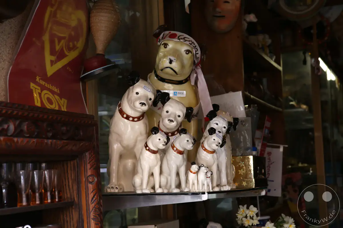 Ein historisches Werbeschild für Spielzeug aus Kurashiki hängt neben einer kunstvollen Sammlung von Nipper-Hundefiguren der Marke RCA Victor auf einem Holzregal im Inneren eines traditionellen japanischen Spielzeugladens.