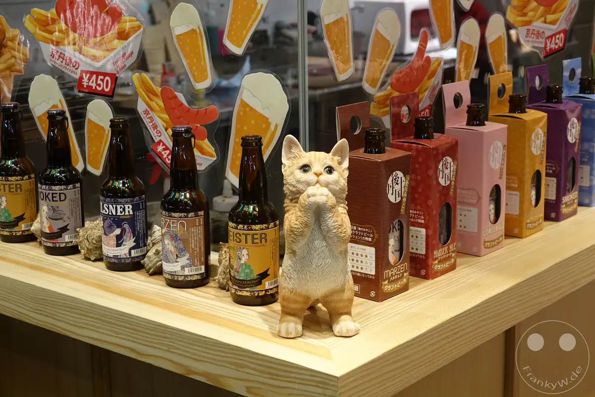 Eine niedliche, aufrecht stehende Katzenfigur schmückt die Schaufensterauslage eines Spezialitätenladens für Craft-Bier in Kyoto, umgeben von verschiedenen Bierflaschen und farbenfrohen Werbeaufstellern.