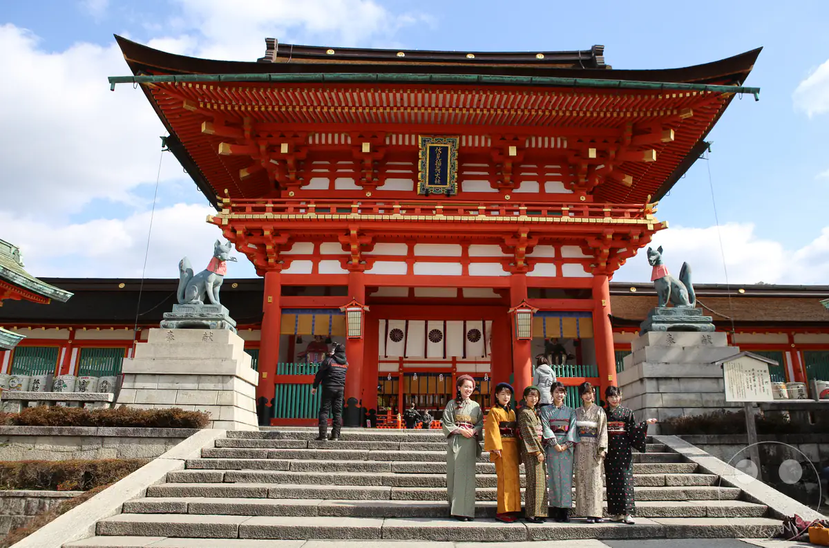 Prächtiges zweistöckiges Romon-Tor am Fushimi-Inari-Taisha-Schrein mit schützenden Kitsune-Fuchsstatuen und einer Gruppe von Frauen in traditionellen Kimonos auf den Steinstufen in Kyoto, Japan.