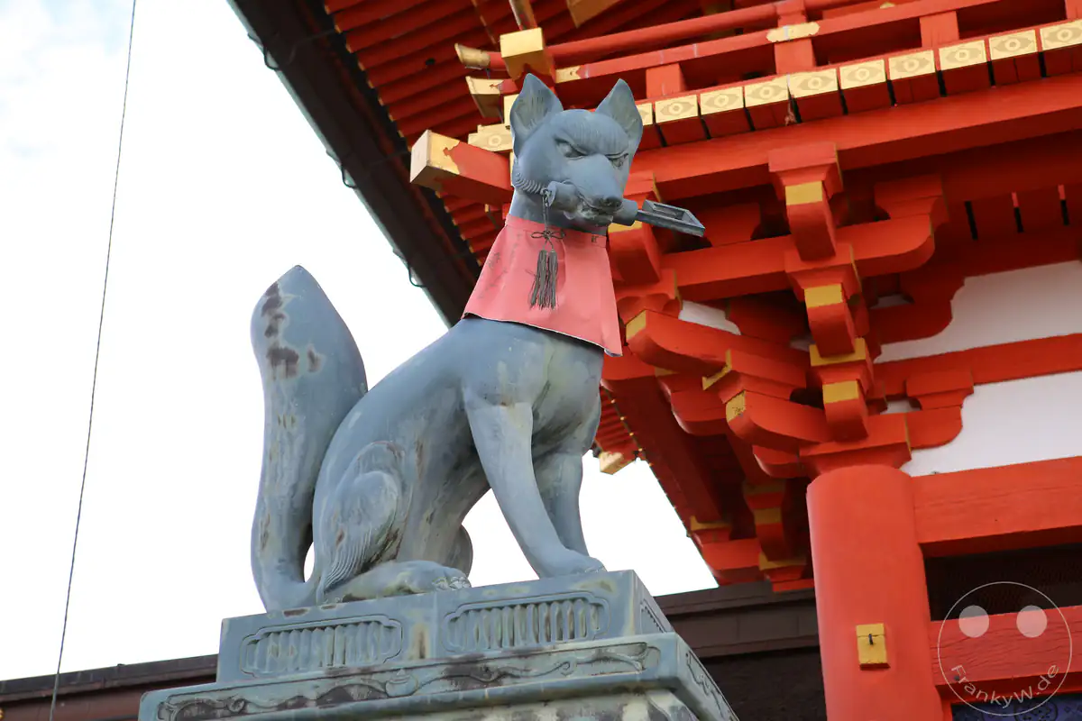 Eine steinerne Kitsune-Fuchsstatue mit einem symbolischen Schlüssel im Maul wacht mit einem roten Lätzchen vor dem prachtvollen, zinnoberroten Romon-Haupttor des Fushimi Inari-Taisha in Kyoto.