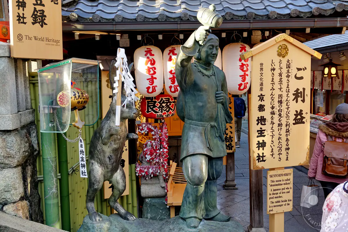 Die bronzene Statue der Shinto-Gottheit Okuninushi mit dem Hasen von Inaba steht neben einer großen hölzernen Informationstafel und traditionellen Papierlaternen am historischen Jishu-Schrein in Kyoto