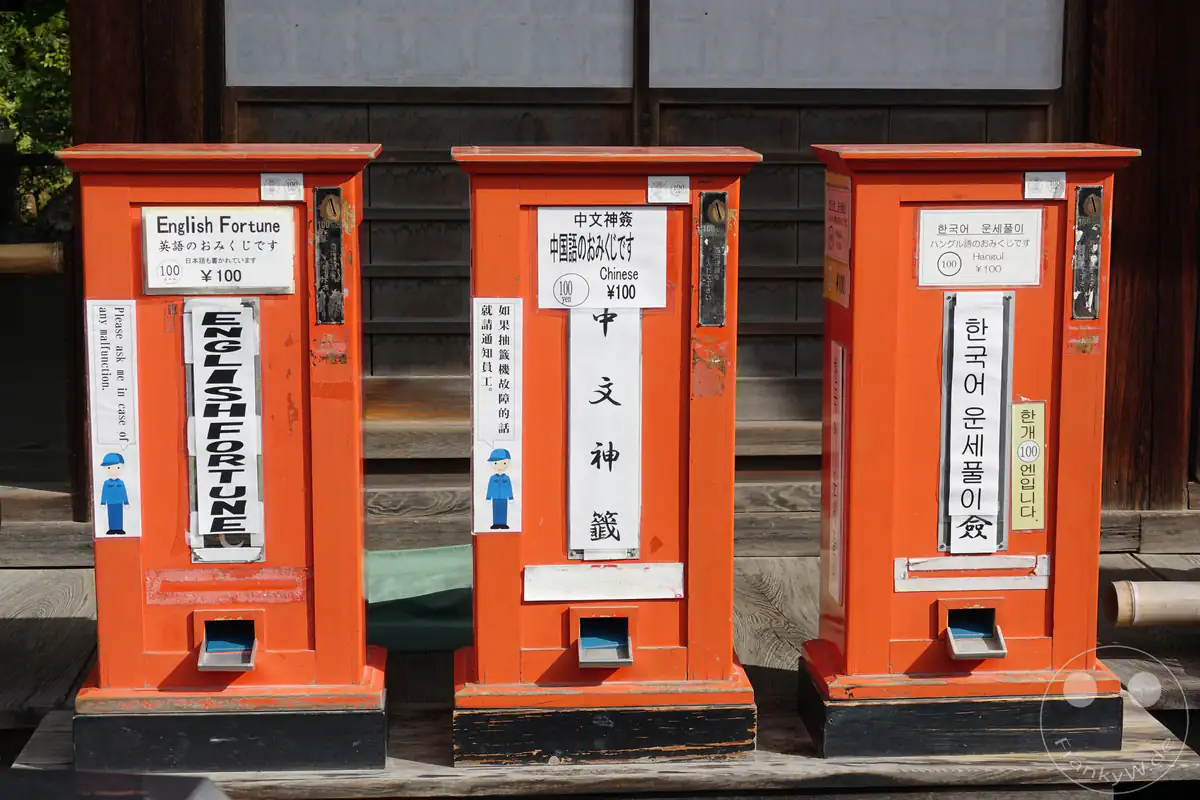 Drei leuchtend orangefarbene Omikuji-Wahrsagungsautomaten für internationale Besucher stehen aufgereiht vor einer Holzwand des Kinkaku-ji Tempels in Kyoto und bieten Orakelzettel auf Englisch, Chinesisch und Koreanisch an