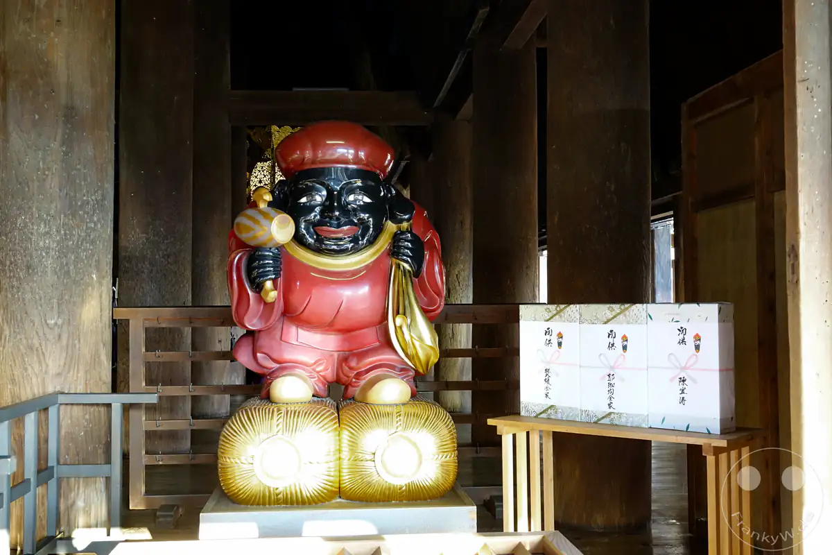 Eine glänzende, farbenfrohe Statue des Glücksgottes Daikokuten mit einem Holzhammer und Reissäcken steht neben verpackten Opfergaben auf einem Holztisch im Inneren des Kiyomizu-dera Tempels in Kyoto.