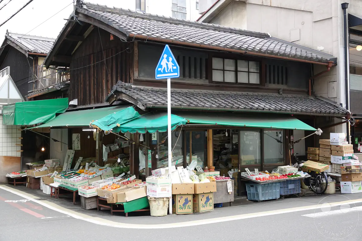 Ein traditioneller japanischer Obst- und Gemüseladen an einer Straßenecke in Kyoto präsentiert frische Waren in Holzkisten und Kartons unter grünen Markisen vor einer historischen Holzfassade mit Ziegeldach.