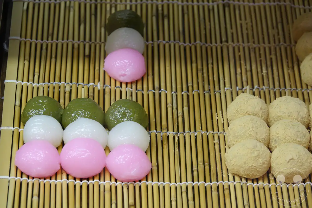 Traditionelle dreifarbige Hanami-Dango-Spieße aus klebrigem Reismehl in Rosa, Weiß und Grün liegen neben Kinako-Mochi auf einer rustikalen Bambusmatte in einer japanischen Süßwarenauslage.