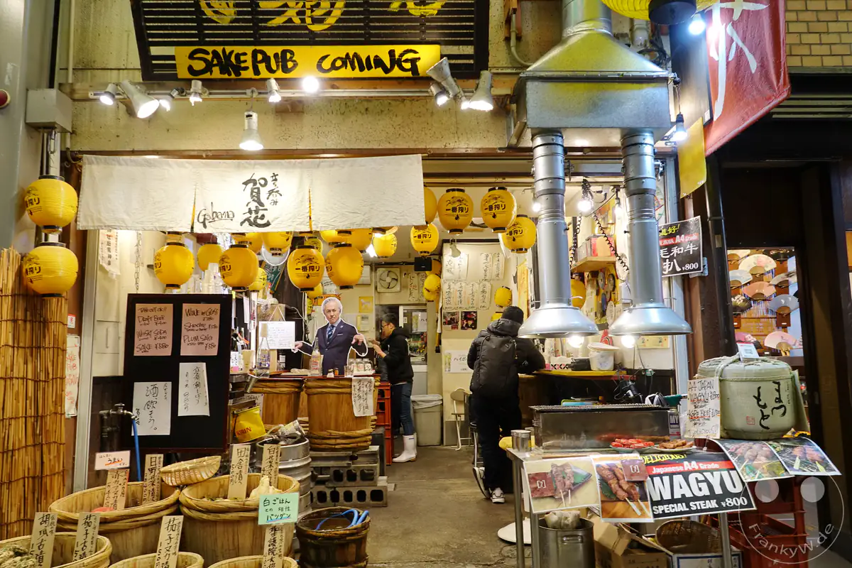 Der einladende Eingang eines traditionellen Sake-Pubs auf dem Nishiki-Markt in Kyoto ist mit gelben Laternen und Noren-Vorhängen geschmückt, während davor ein Grillstand frisches Wagyu-Steak anbietet.