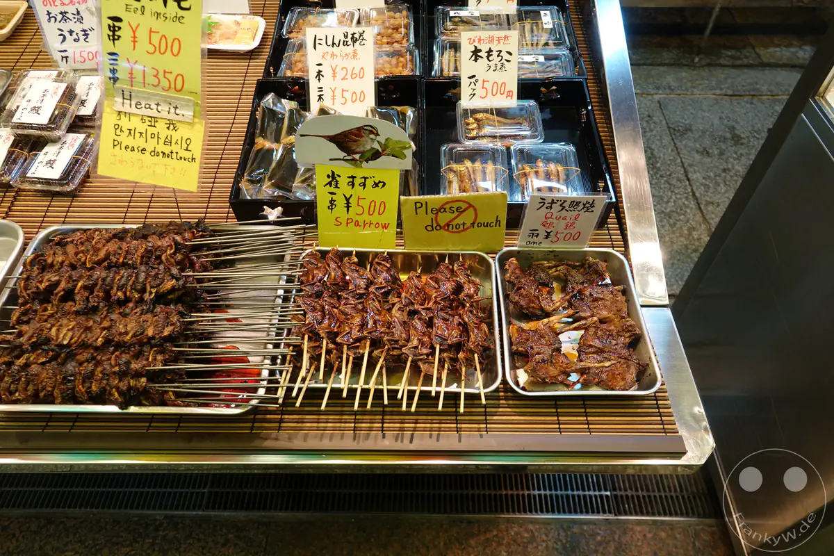 Gegrillte Spieße von Spatz und Wachtel sowie Aalkonfekt liegen neben handgeschriebenen, mehrsprachigen Preisschildern in der Auslage eines traditionellen Marktstandes in Kyoto.