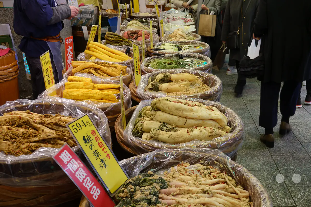 Eine lange Reihe traditioneller Holzfässer präsentiert eine große Auswahl an Tsukemono, japanischem eingelegtem Gemüse wie Daikon-Rettich, mit gelben Preisschildern auf dem Nishiki-Markt in Kyoto.