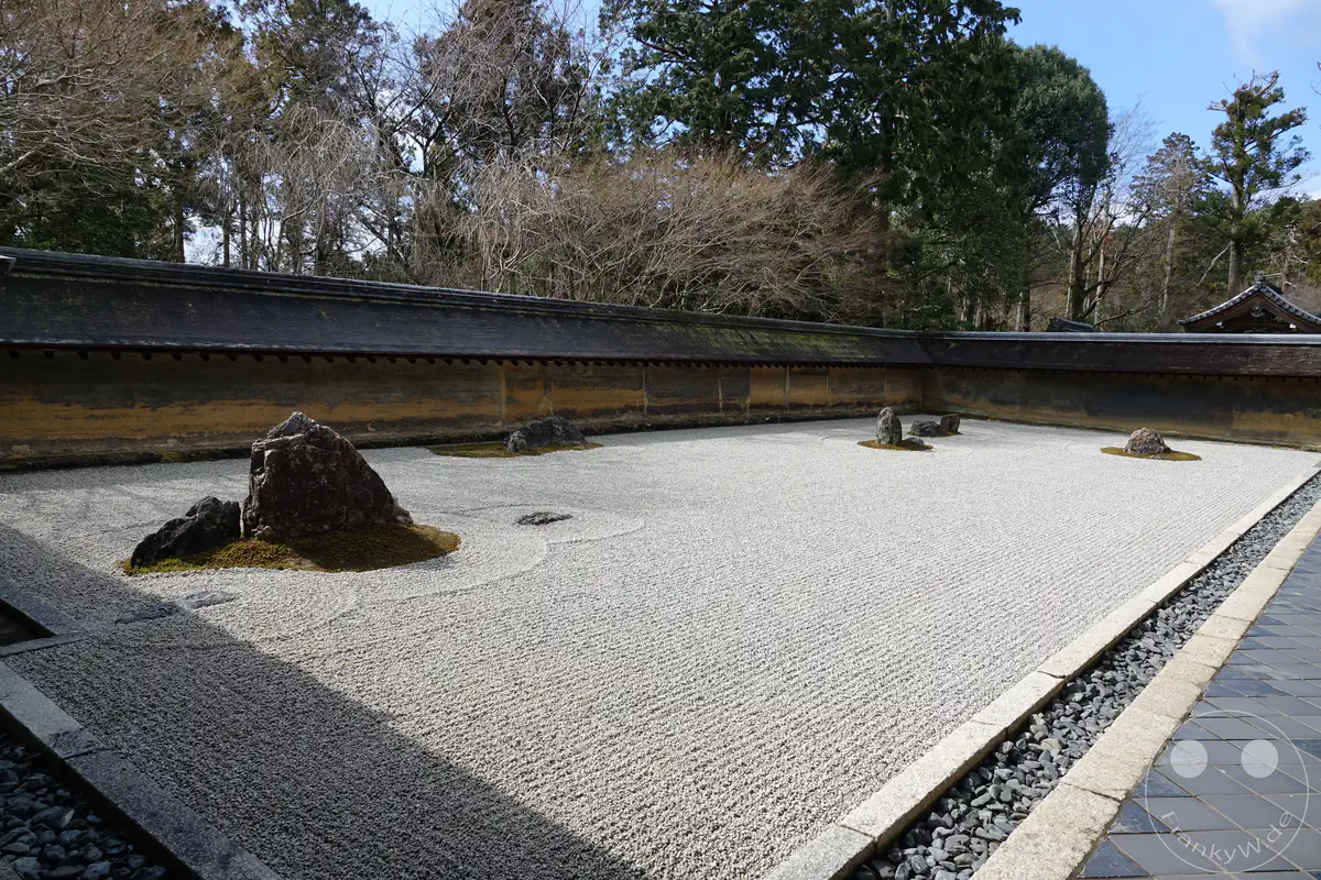 Der berühmte, von einer niedrigen Lehmmauer umgebene Zen-Steingarten des Ryoan-ji Tempels in Kyoto zeigt sorgfältig geharkten weißen Kies und kunstvoll platzierte Felsformationen unter blauem Himmel.