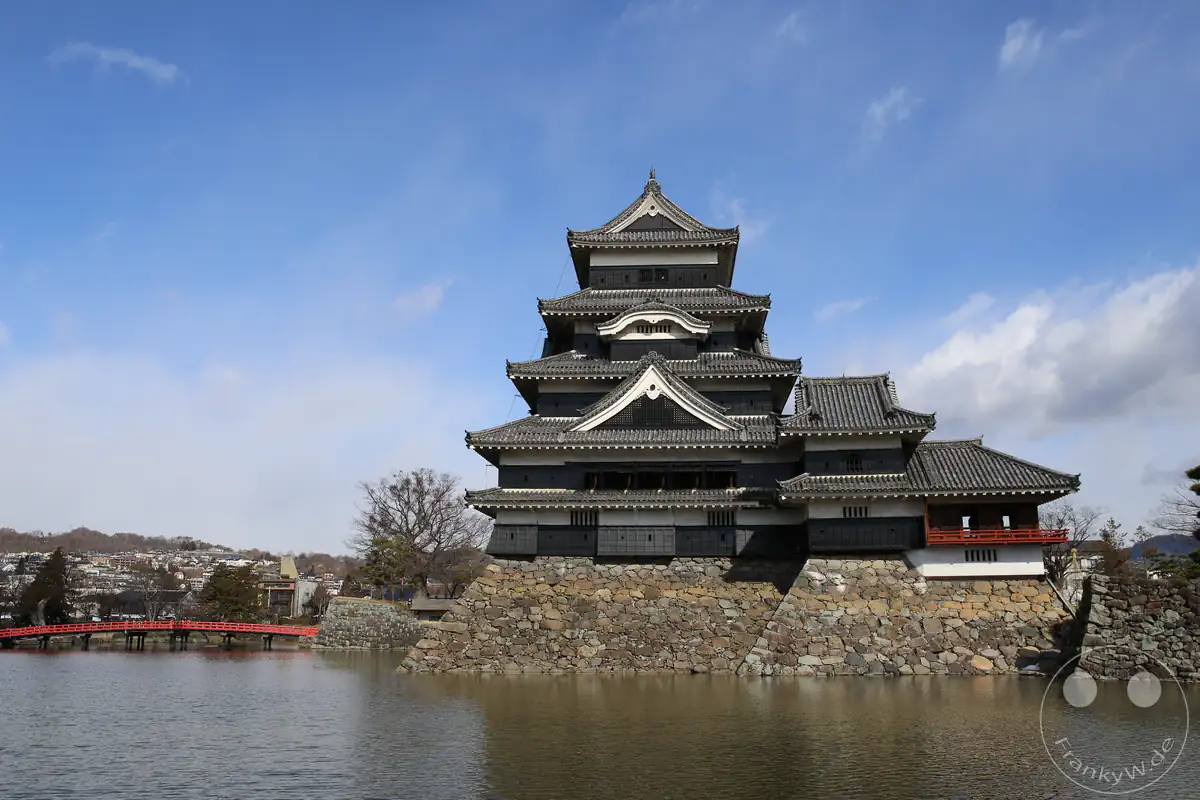 Die historische Burg Matsumoto mit ihrer charakteristischen schwarzen Holzfassade, den mehrstöckigen Dächern und einer roten Holzbrücke spiegelt sich im umliegenden Wassergraben unter blauem Himmel in Matsumoto in der Präfektur Nagano, Japan.