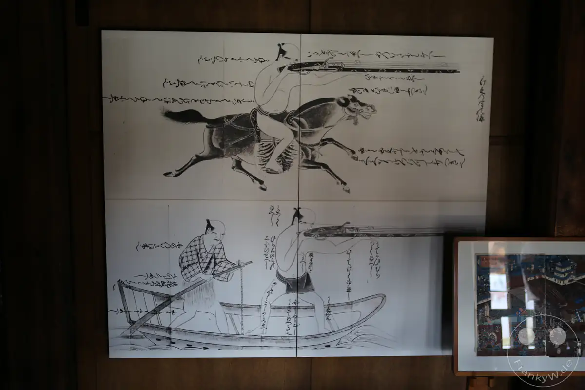 Eine Schautafel im Inneren der Burg Matsumoto zeigt historische Tuschezeichnungen und japanische Schriftzeichen, die den Umgang mit Luntenschlossgewehren (Tanegashima) zu Pferd und vom Boot aus darstellen, in Matsumoto in der Präfektur Nagano, Japan.