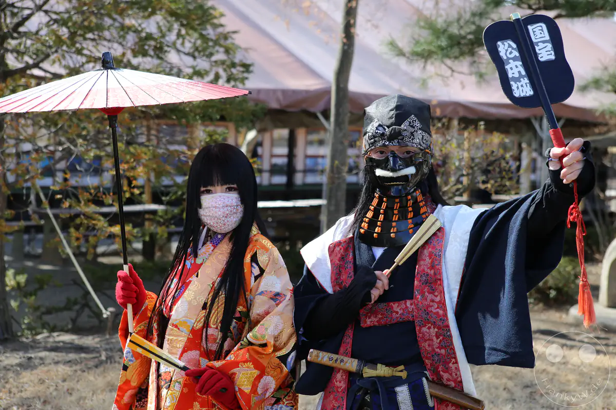 Zwei Schausteller in aufwendigen historischen Gewändern posieren im Park der Burg Matsumoto, wobei die Frau einen prachtvollen Kimono mit rotem Sonnenschirm trägt und der Mann eine traditionelle Rüstung mit Gesichtsmaske (Menpo) präsentiert in Matsumoto in der Präfektur Nagano, Japan.