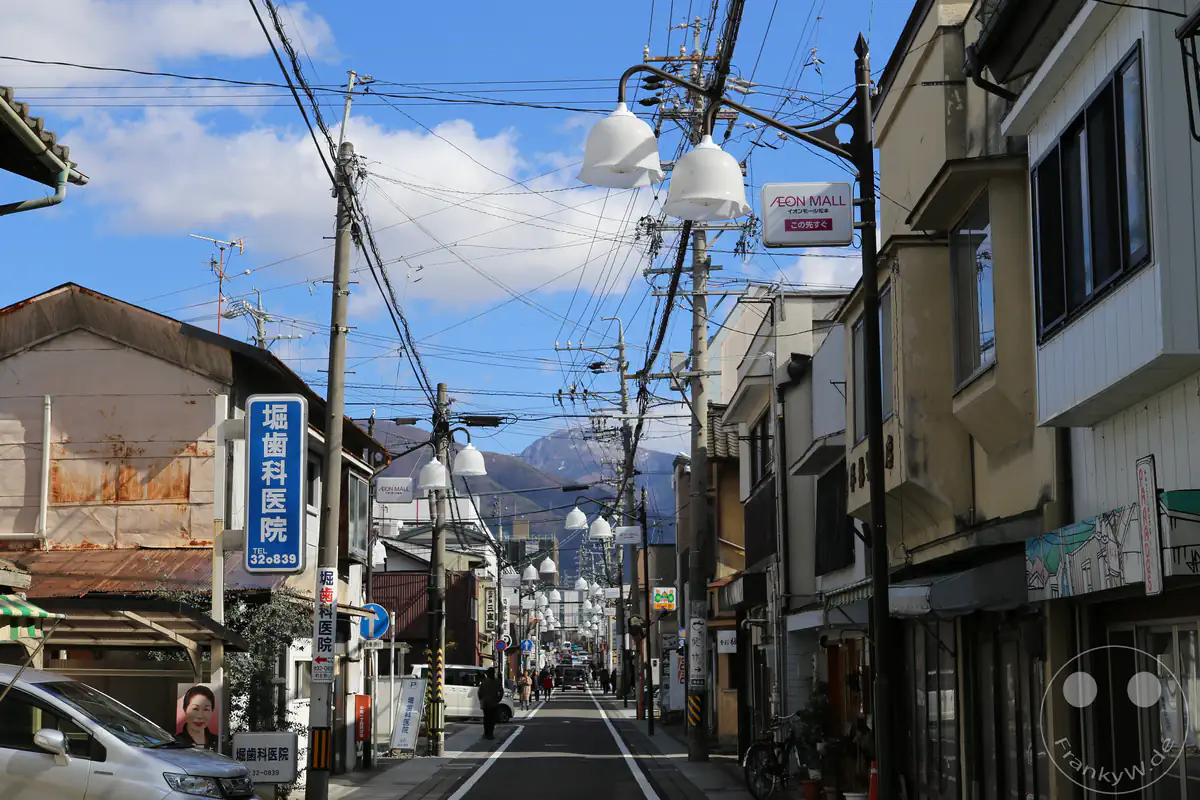 Blick durch eine traditionelle Einkaufsstraße in Matsumoto mit charakteristischen weißen Gebäuden im Lagerhaus-Stil (Kura), markanten Straßenlaternen, Überlandleitungen und den schneebedeckten Gipfeln der Japanischen Alpen im Hintergrund in der Präfektur Nagano, Japan.