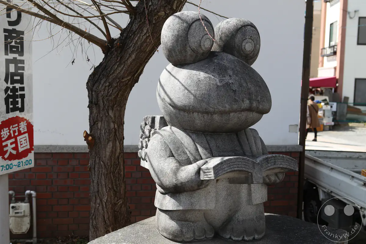 Eine humorvolle Steinstatue eines Frosches in der Pose des Gelehrten Ninomiya Sontoku, der beim Gehen ein Buch liest und Reisig auf dem Rücken trägt, steht in der historischen Nawate-Straße (Frosch-Straße) in Matsumoto in der Präfektur Nagano, Japan.