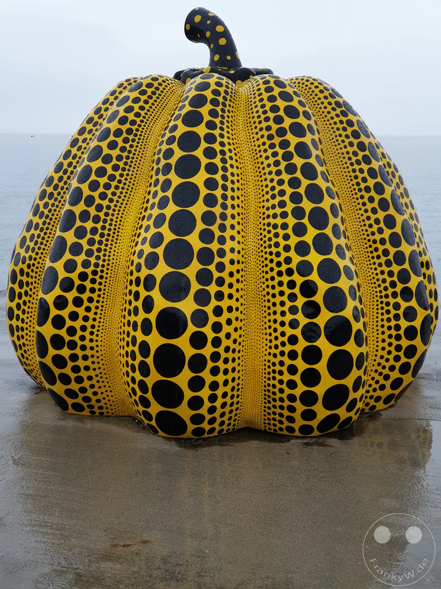 Die ikonische gelbe Kürbis-Skulptur mit schwarzen Punkten der Künstlerin Yayoi Kusama steht an der Küste der Kunstinsel Naoshima direkt am Meer unter einem wolkenverhangenen Himmel.