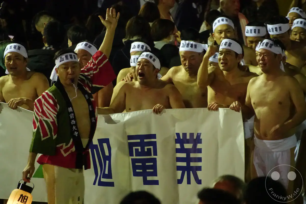 Teilnehmer des Hadaka-Matsuri-Festivals in Fundoshi-Lendentüchern und Stirnbändern rufen lautstark Parolen, während ein Mann in einer Happi-Jacke die Gruppe am Saidai-ji Kannon-in Tempel in Okayama anführt.