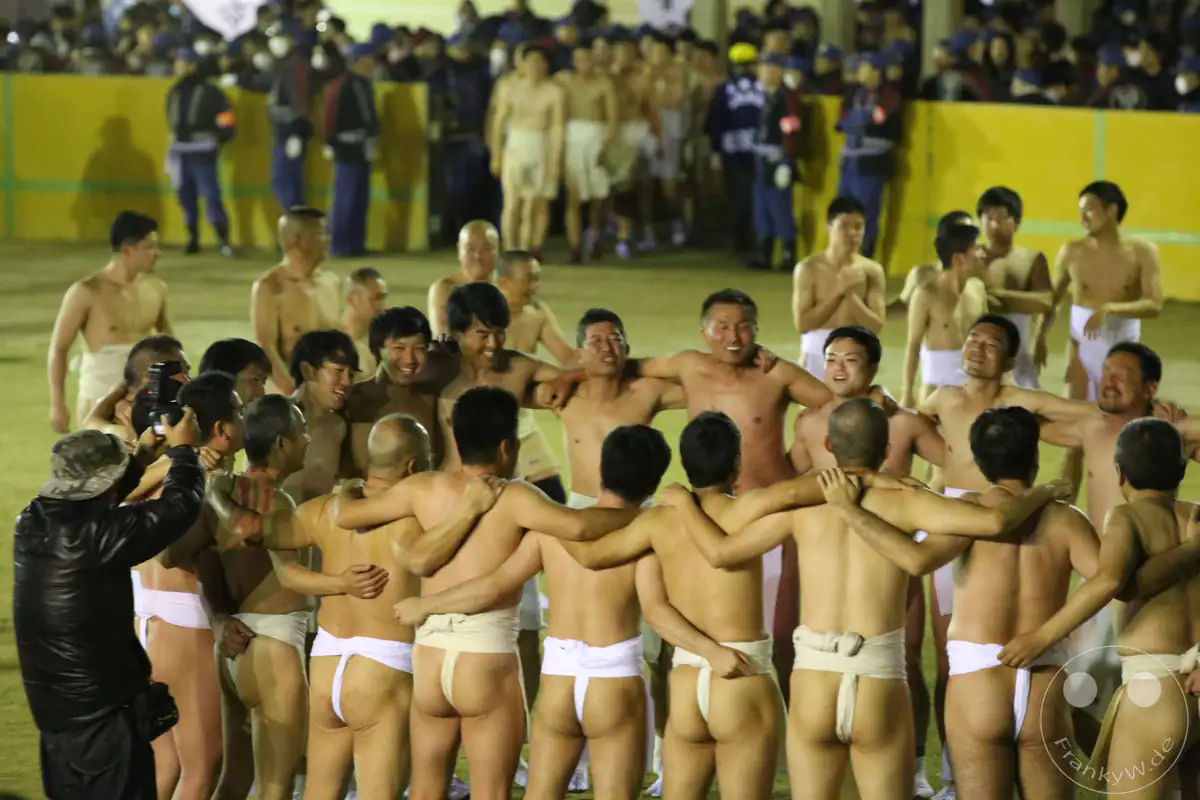 Eine Gruppe nackter Männer in traditionellen Fundoshi-Lendentüchern bildet Arm in Arm einen engen Kreis, während ein Fotograf die emotionale Kameradschaft beim Hadaka-Matsuri in Okayama festhält.