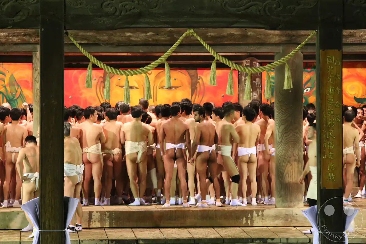 Hunderte nackte Männer in weißen Fundoshi-Lendentüchern drängen sich in einer hölzernen Tempelhalle vor einer großen, farbenfrohen Buddha-Wandmalerei beim traditionellen Hadaka-Matsuri in Okayama.