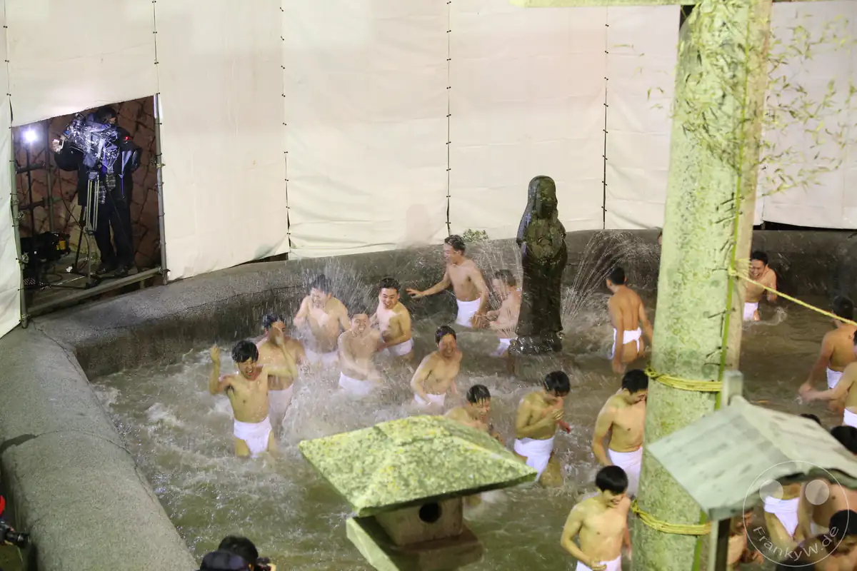 Nackte Männer in weißen Fundoshi-Lendentüchern waten johlend durch ein rituelles Reinigungsbecken mit einer Kannon-Statue während des traditionellen Hadaka-Matsuri am Tempel Saidai-ji in Okayama