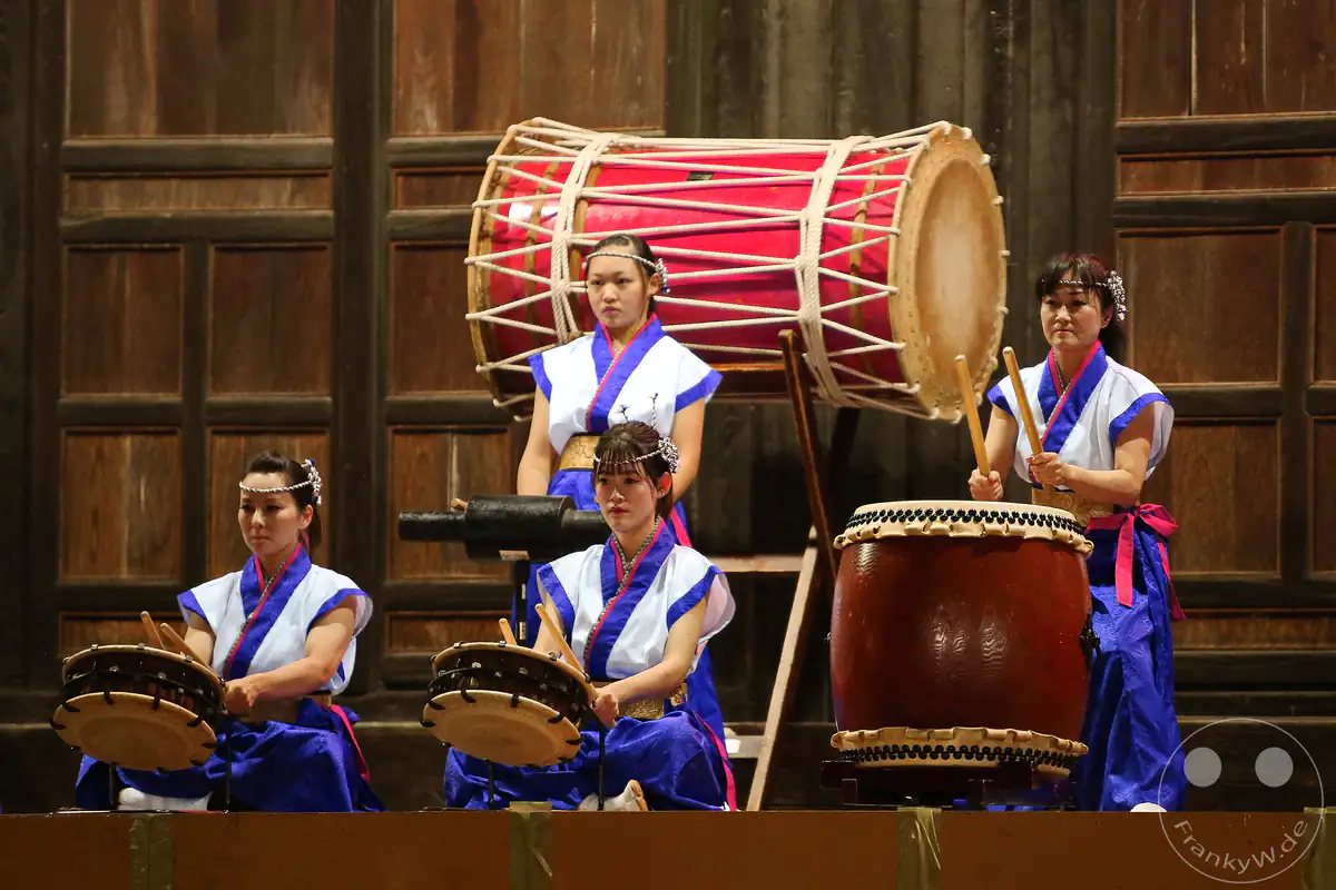 Vier japanische Musikerinnen präsentieren ein kraftvolles Taiko-Trommelkonzert in traditionellen Gewändern vor einer historischen Holzwand beim Hadaka-Matsuri im Saidai-ji Kannon-in Tempel in Okayama.
