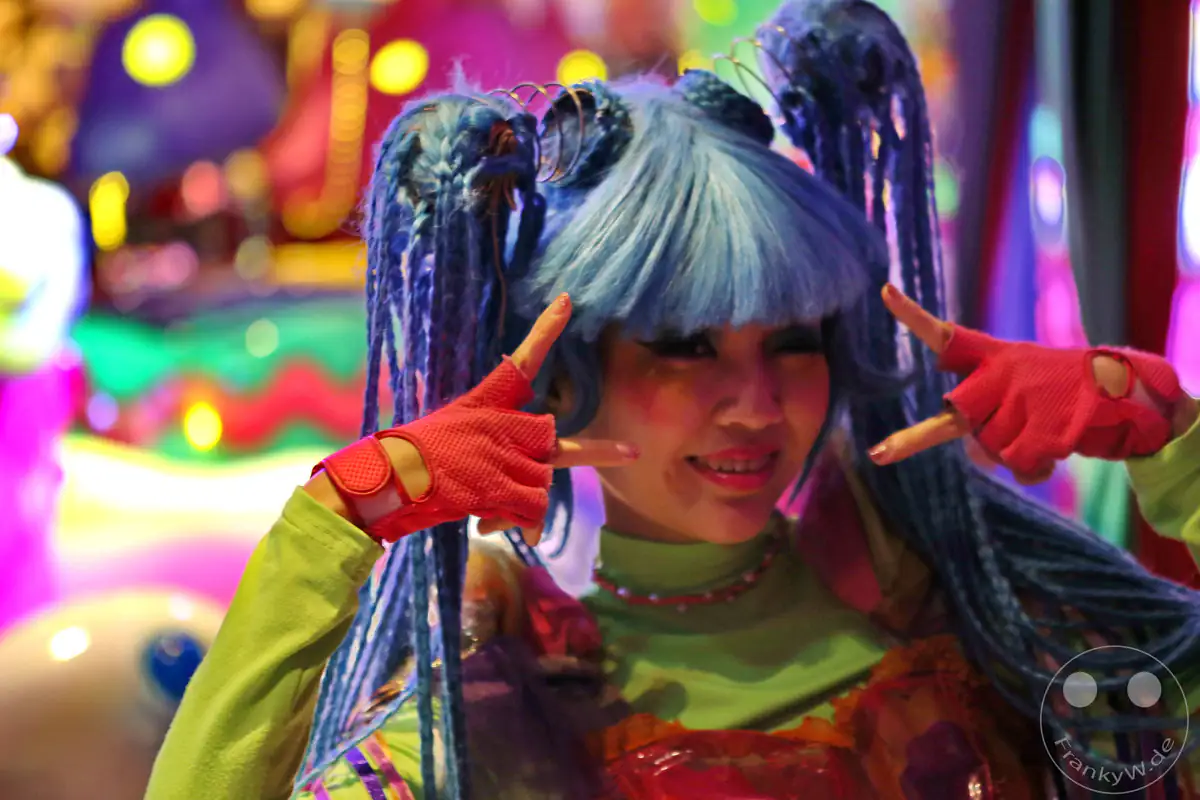 Eine Darstellerin im Kawaii-Monster-Cafe im Stadtteil Harajuku posiert in einem bunten Kostüm mit leuchtend blauer Perücke, geflochtenen Zöpfen und pinkfarbenen fingerlosen Handschuhen mit dem Peace-Zeichen vor einem farbenfrohen, hell erleuchteten Hintergrund in Tokio, Japan