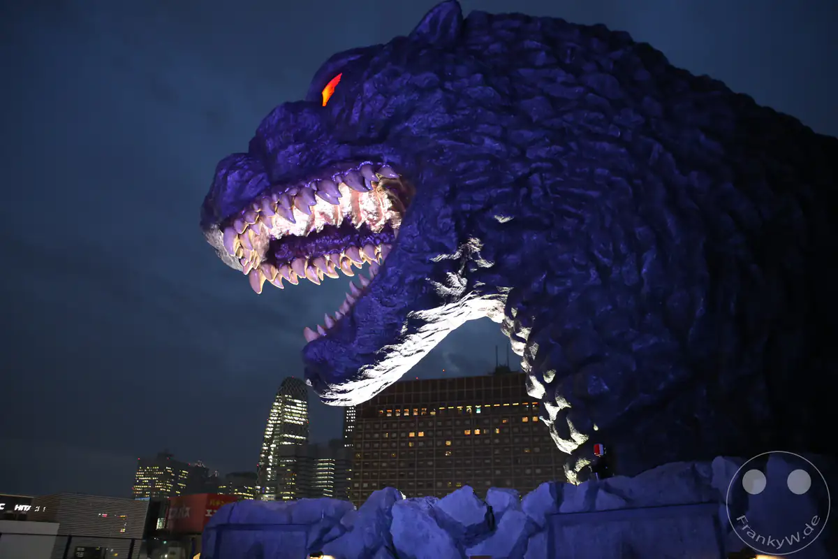 Der riesige, lebensgroße Godzilla-Kopf auf der Dachterrasse des Shinjuku-Toho-Gebäudes brüllt bei Nacht mit beleuchtetem, offenem Maul und rot leuchtenden Augen über der Skyline des Amüsierviertels Kabukicho im Stadtteil Shinjuku in Tokio, Japan.