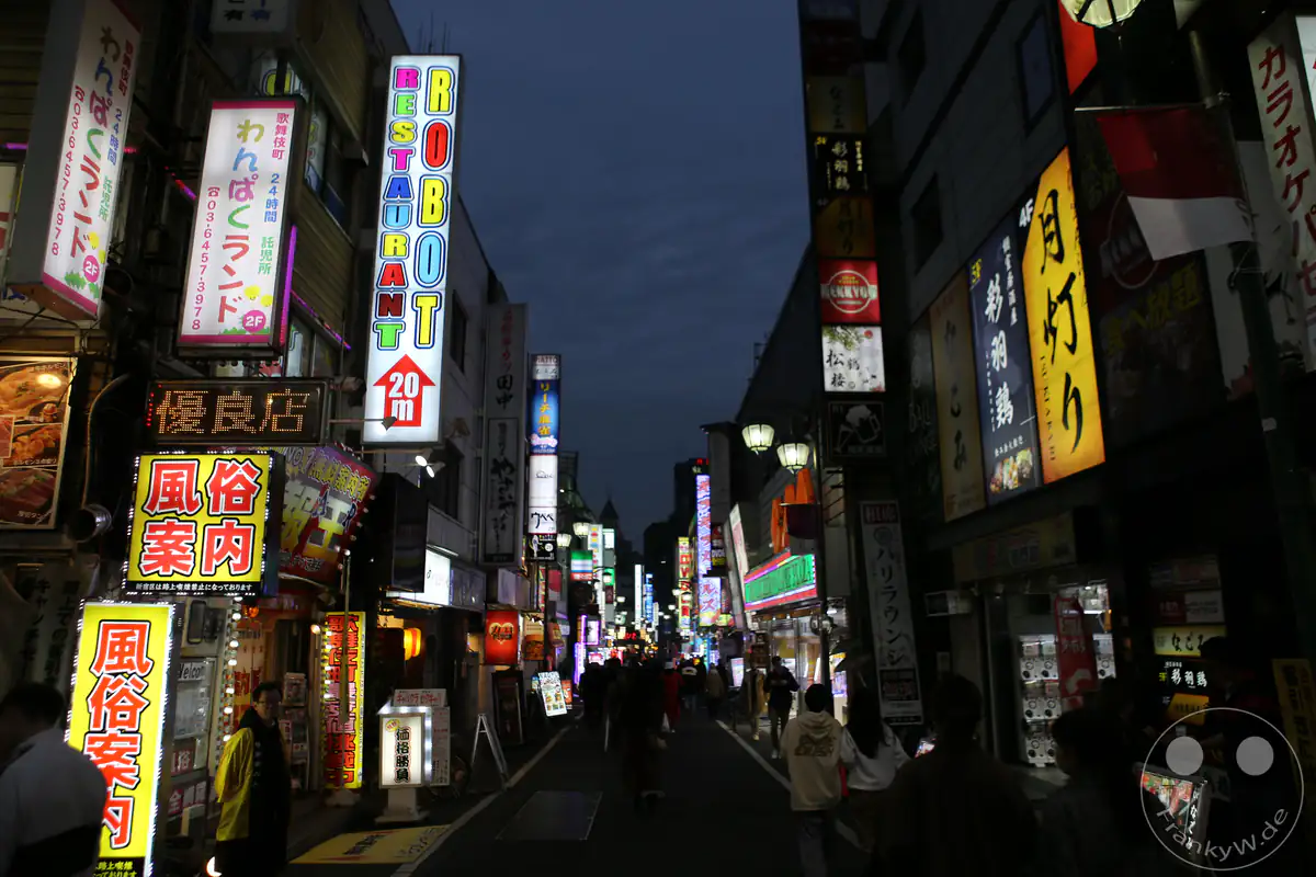 Blick durch eine belebte, eng bebaute Straße im Amüsierviertel Kabukicho bei Nacht mit leuchtenden Neon-Reklamen, japanischen Schriftzeichen und dem markanten Hinweisschild zum berühmten Robot Restaurant im Stadtteil Shinjuku in Tokio, Japan.