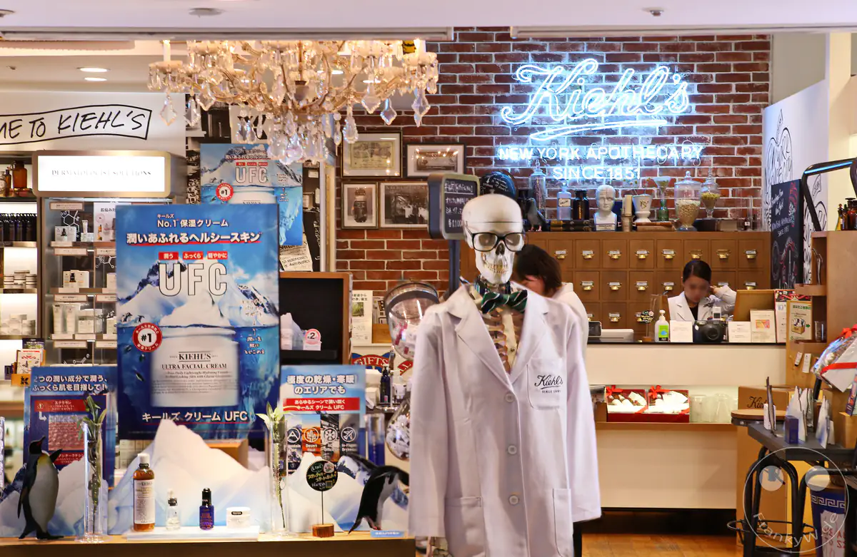 Ein markantes Kiehl's Kosmetikgeschäft im Stil einer New Yorker Apotheke mit einer dekorativen Skelettfigur im weißen Labormantel und Brille, einer Backsteinwand mit Neon-Schriftzug und großen Werbeaufstellern in Tokio, Japan.
