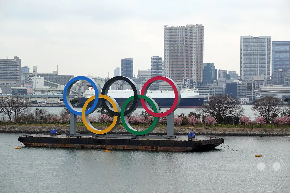 Das riesige schwimmende Monument der fünf Olympischen Ringe steht auf einer Transportbarke im Wasser des Odaiba Marine Parks vor der modernen Skyline der Tokyo-Bay unter einem hellen, bewölkten Himmel in Tokio, Japan.