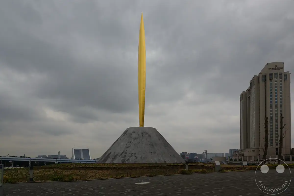 Die goldene Skulptur der Statue der Freiheit (Flame of Liberty) des französischen Bildhauers Marc Couturier ragt auf einem grauen, pyramidenartigen Betonsockel vor der Kulisse des Hotels Grand Nikko Tokyo Daiba unter einem tristen, wolkenverhangenen Himmel empor auf der künstlichen Insel Odaiba im Stadtteil Koto in Tokio, Japan.