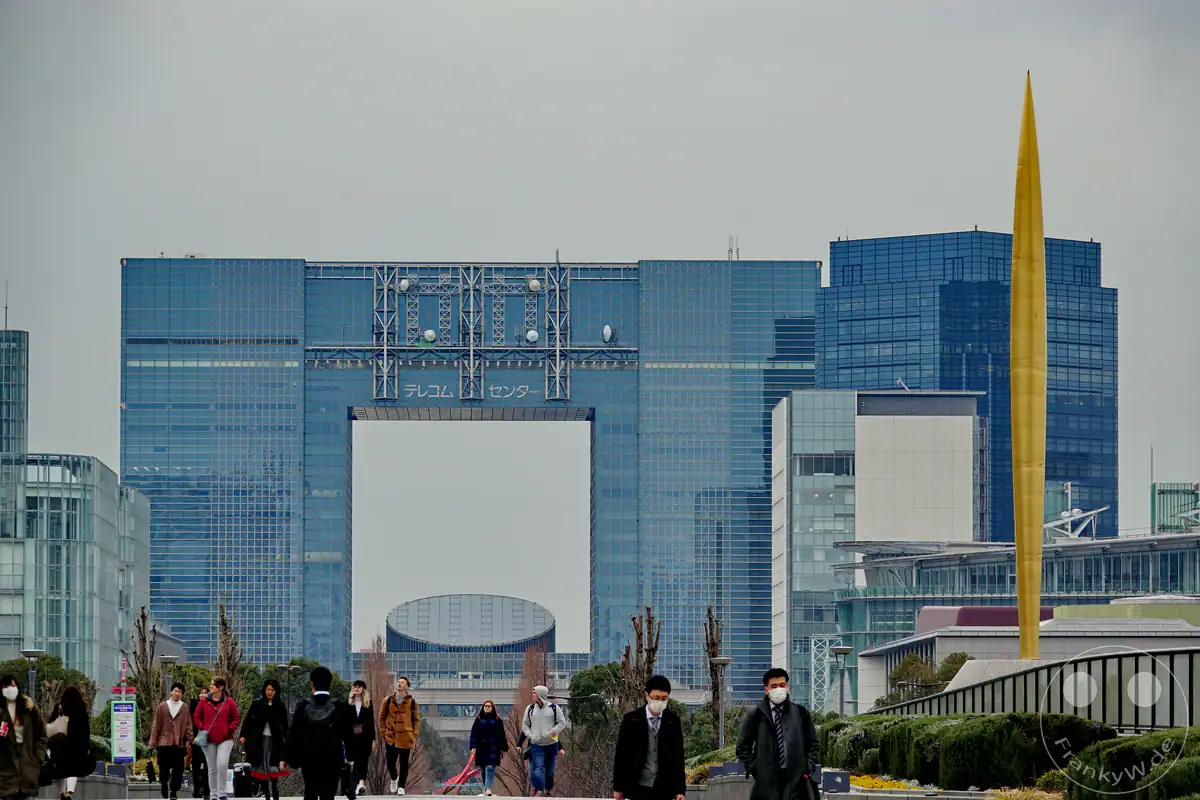 Das markante, bogenförmige Telecom-Center-Gebäude mit seiner bläulichen Glasfassade bildet die Kulisse für Passanten auf der Symbol-Promenade-Park-Flaniermeile neben der goldenen Flamme-der-Freiheit-Skulptur auf der künstlichen Insel Odaiba im Stadtteil Koto in Tokio, Japan.