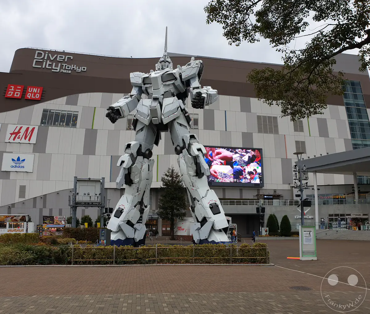 Die lebensgroße, weiße Statue des RX-0 Unicorn Gundam steht auf einem gepflasterten Vorplatz vor der modernen Fassade des Einkaufszentrums DiverCity Tokyo Plaza mit Logos bekannter Modemarken und einem großen Bildschirm auf der künstlichen Insel Odaiba im Stadtteil Koto in Tokio, Japan.