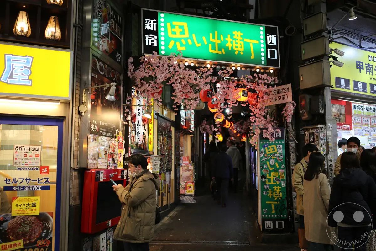 Das markante grüne Neonschild markiert den westlichen Eingang zur historischen Omoide-Yokocho-Gasse nahe dem Bahnhof Shinjuku, dekoriert mit künstlichen Kirschblütenzweigen und Laternen über den Köpfen von Passanten im Stadtteil Shinjuku in Tokio, Japan.